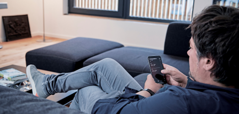 Mann auf Couch, mit Handy in der Hand worauf Dimplex Home App zusehen ist